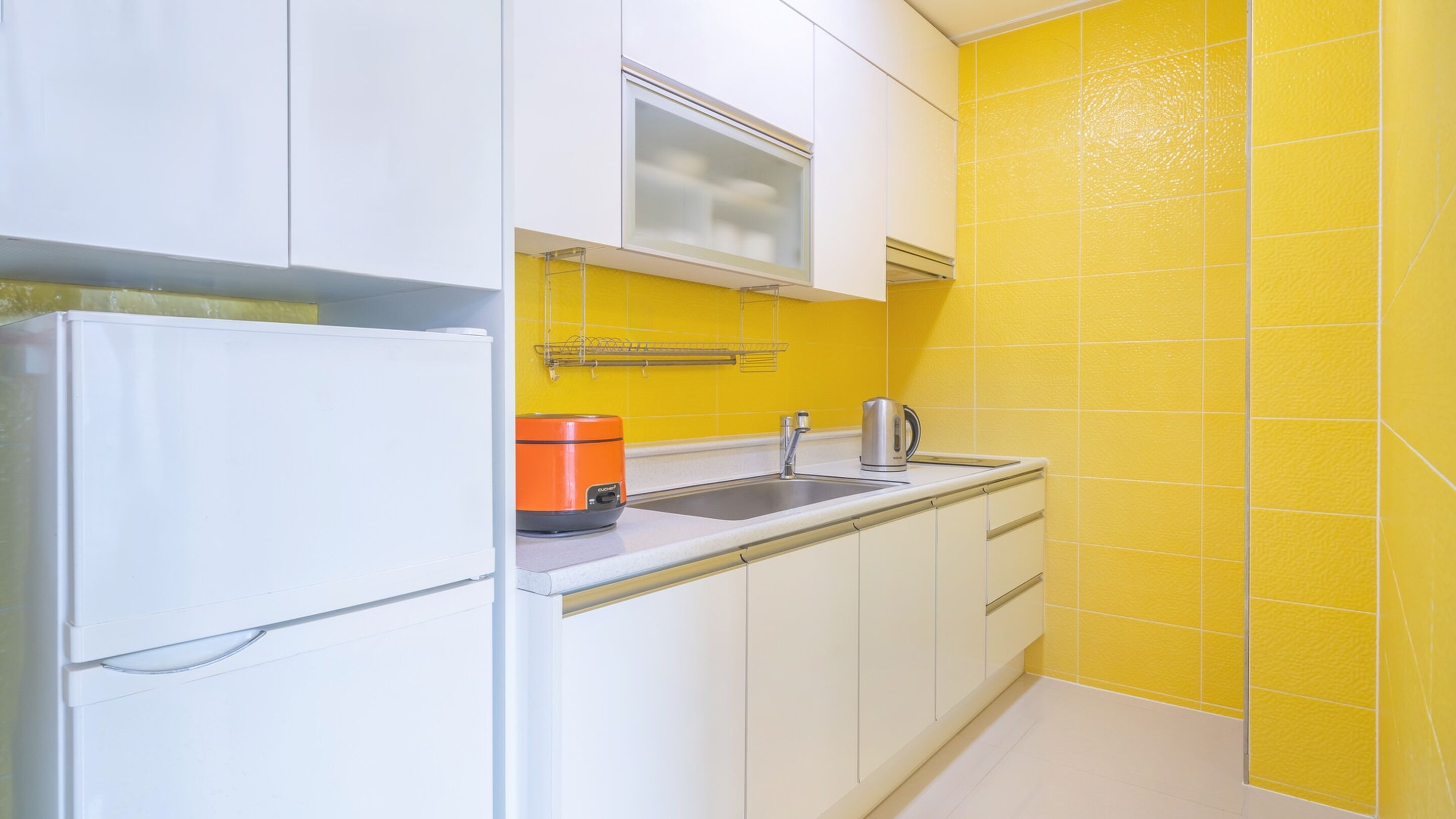 A kitchen with white cabinets, a yellow tiled backsplash, a stainless steel sink, and a kettle.
