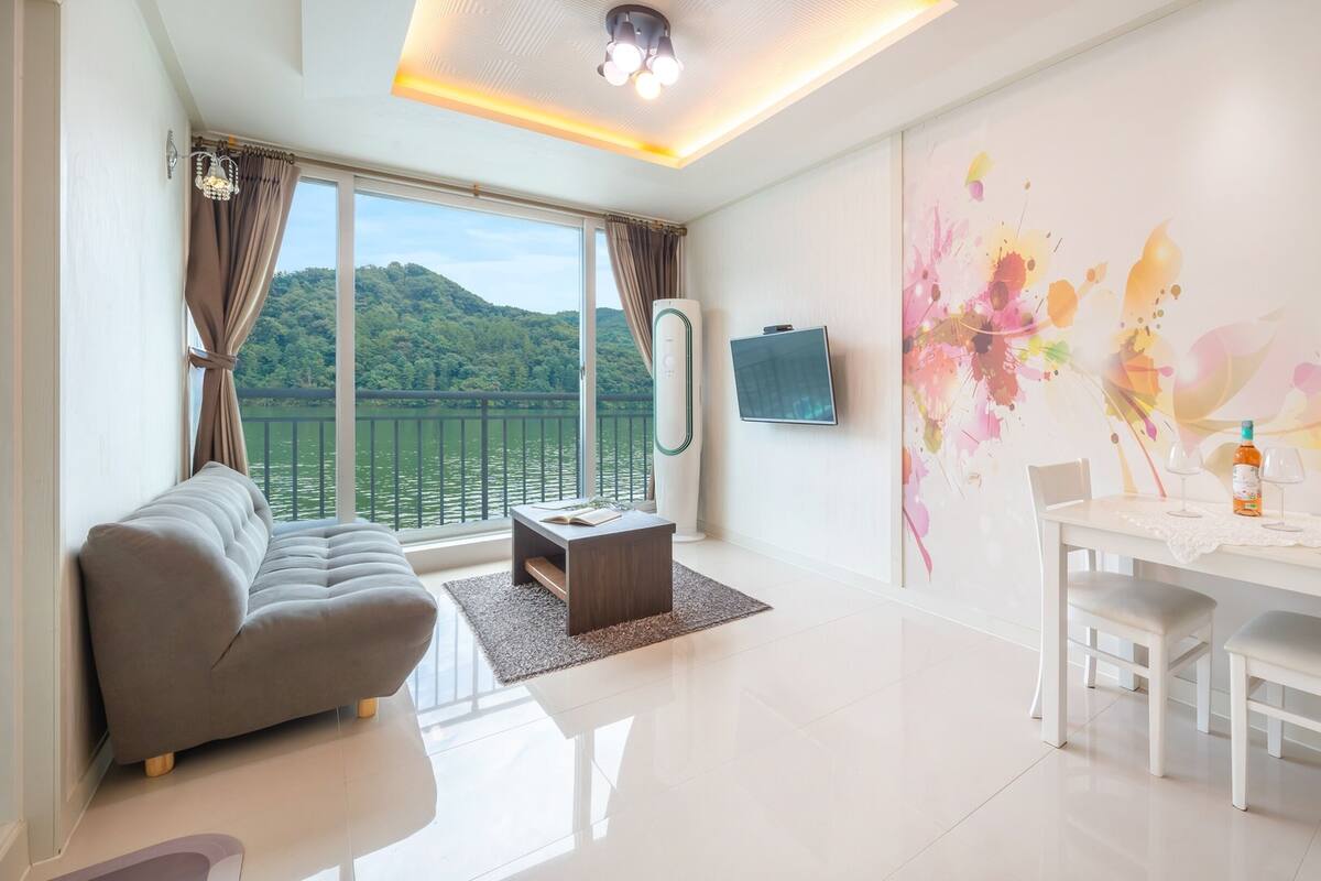 A modern living room with a sofa, dining table, and a view of a lake through large windows.