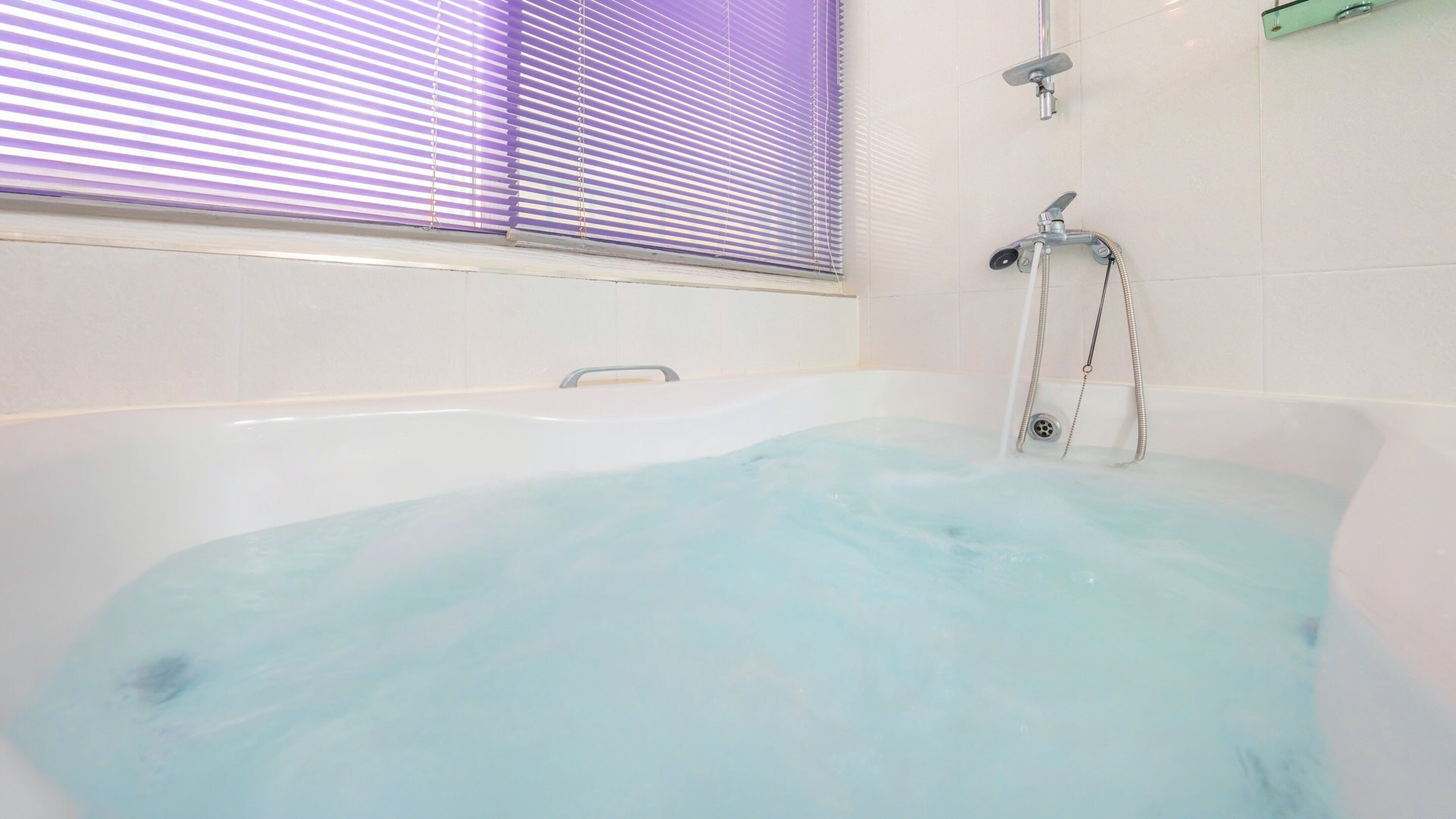 Una bañera con agua corriente, un cabezal de ducha y una ventana con persianas.