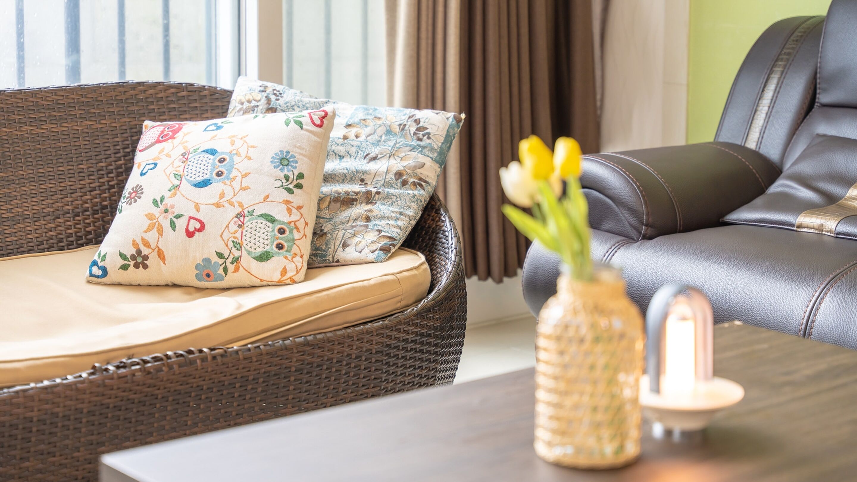 A wicker sofa with floral cushions, a vase with yellow flowers, and a leather armchair.