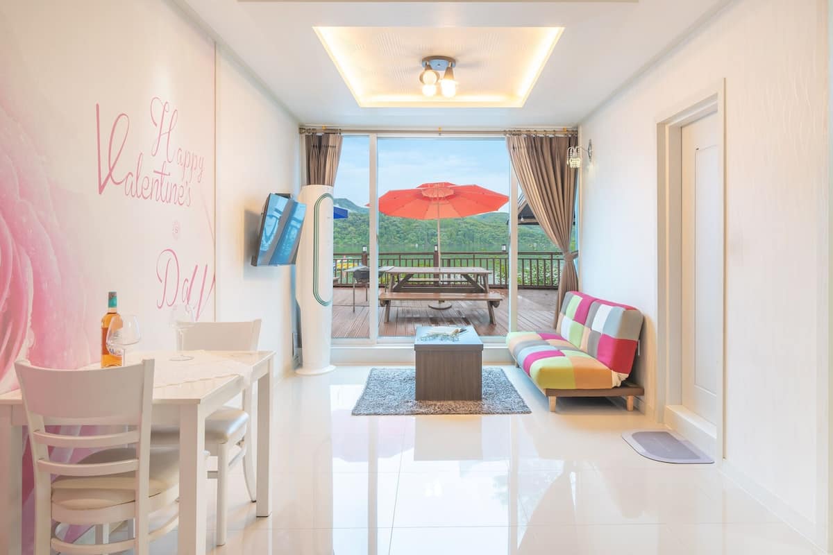 A modern living room with a white sofa, a dining table with chairs, a flat-screen TV mounted on the wall, and a large window with a view of greenery.