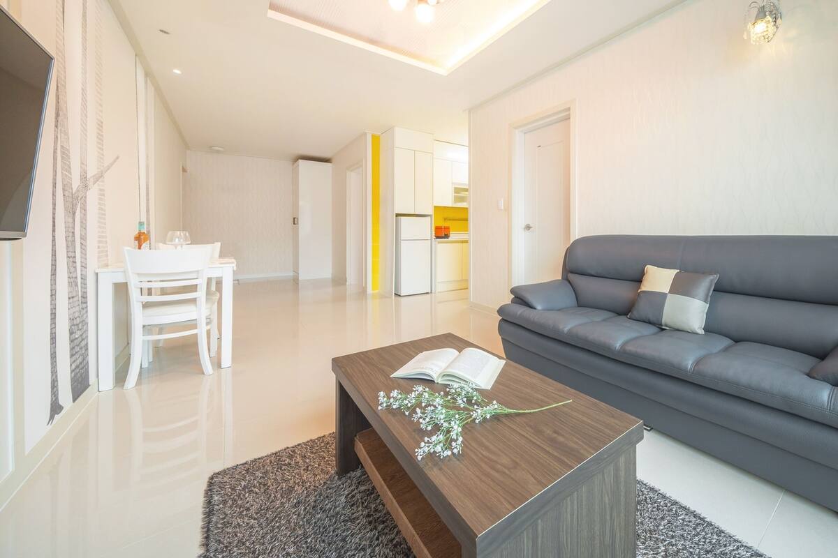 A modern living room with a grey sofa, a wooden coffee table with a book and flowers, a white dining table with chairs, and a kitchen in the background.