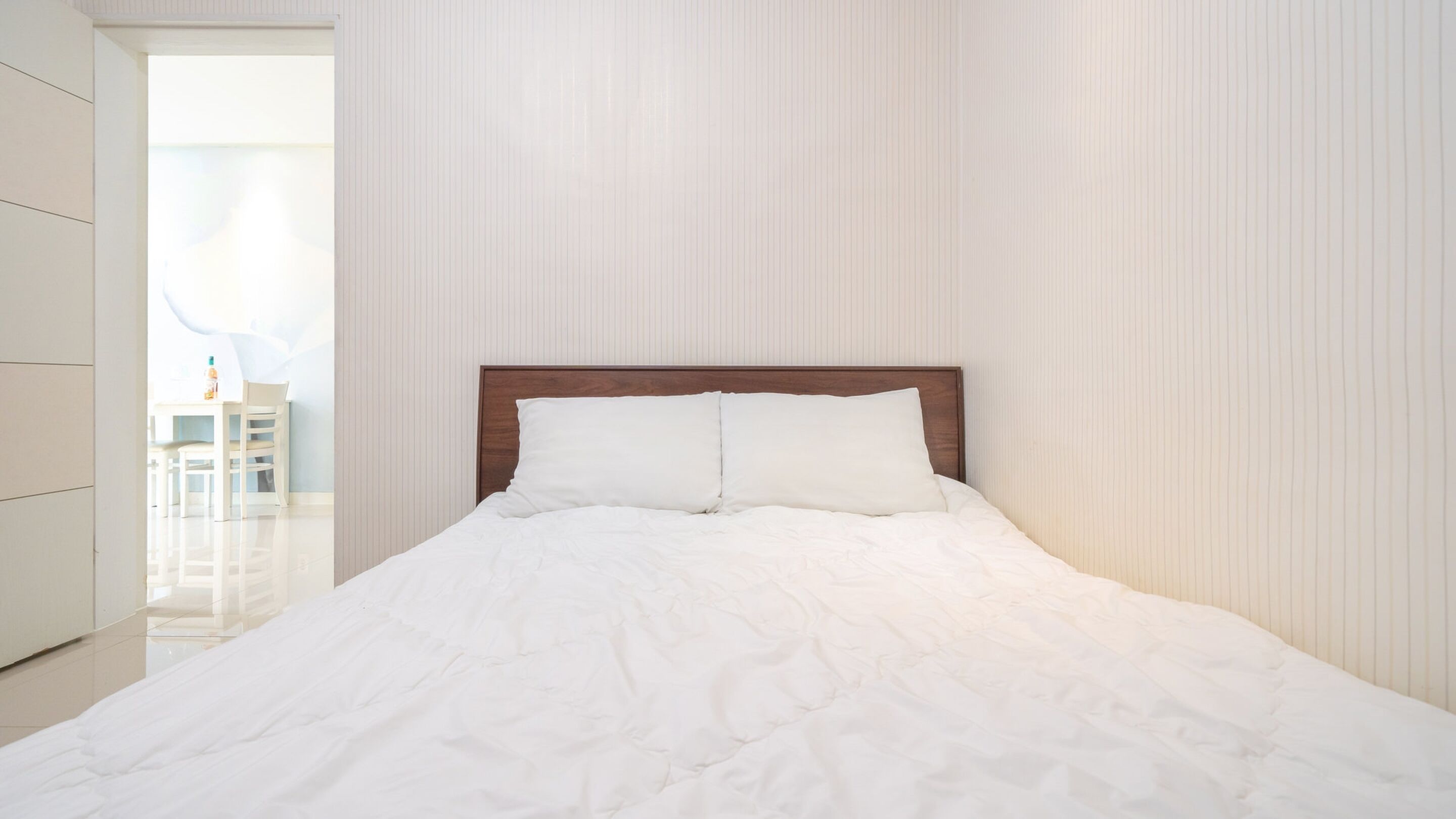 Una cama bien tendida con cabecera de madera en una habitación con paredes blancas y un suelo claro y limpio.