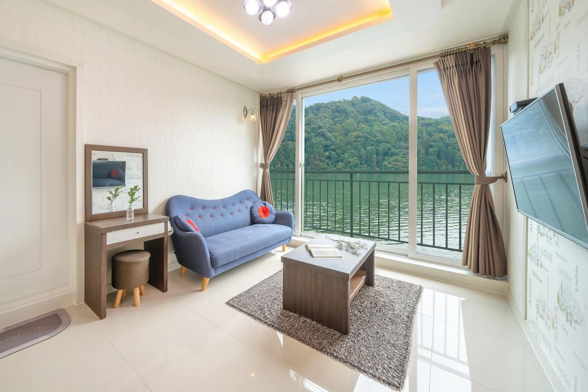A modern living room with a sofa, a small desk, a TV, and a large window with a view of a lake and forest.