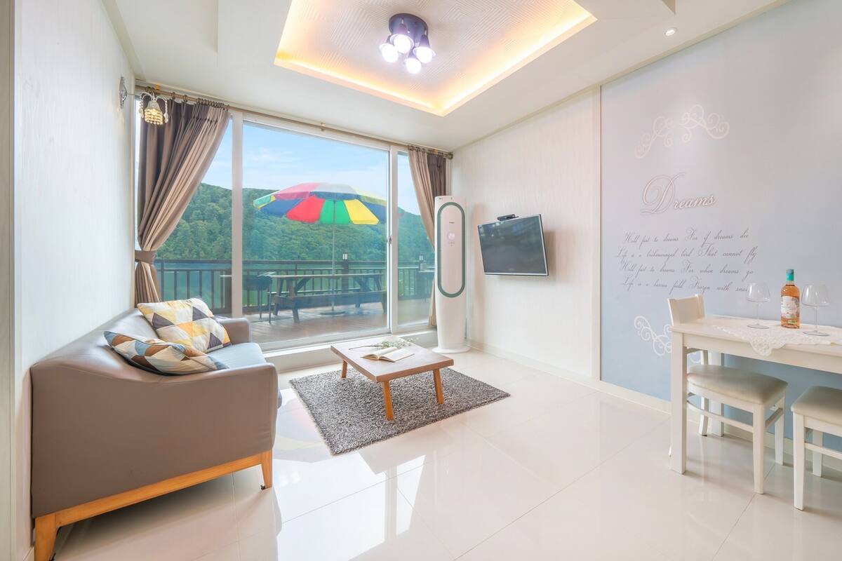 A modern living room with a sofa, coffee table, and dining table. A large window offers a view of a balcony with a colorful umbrella.