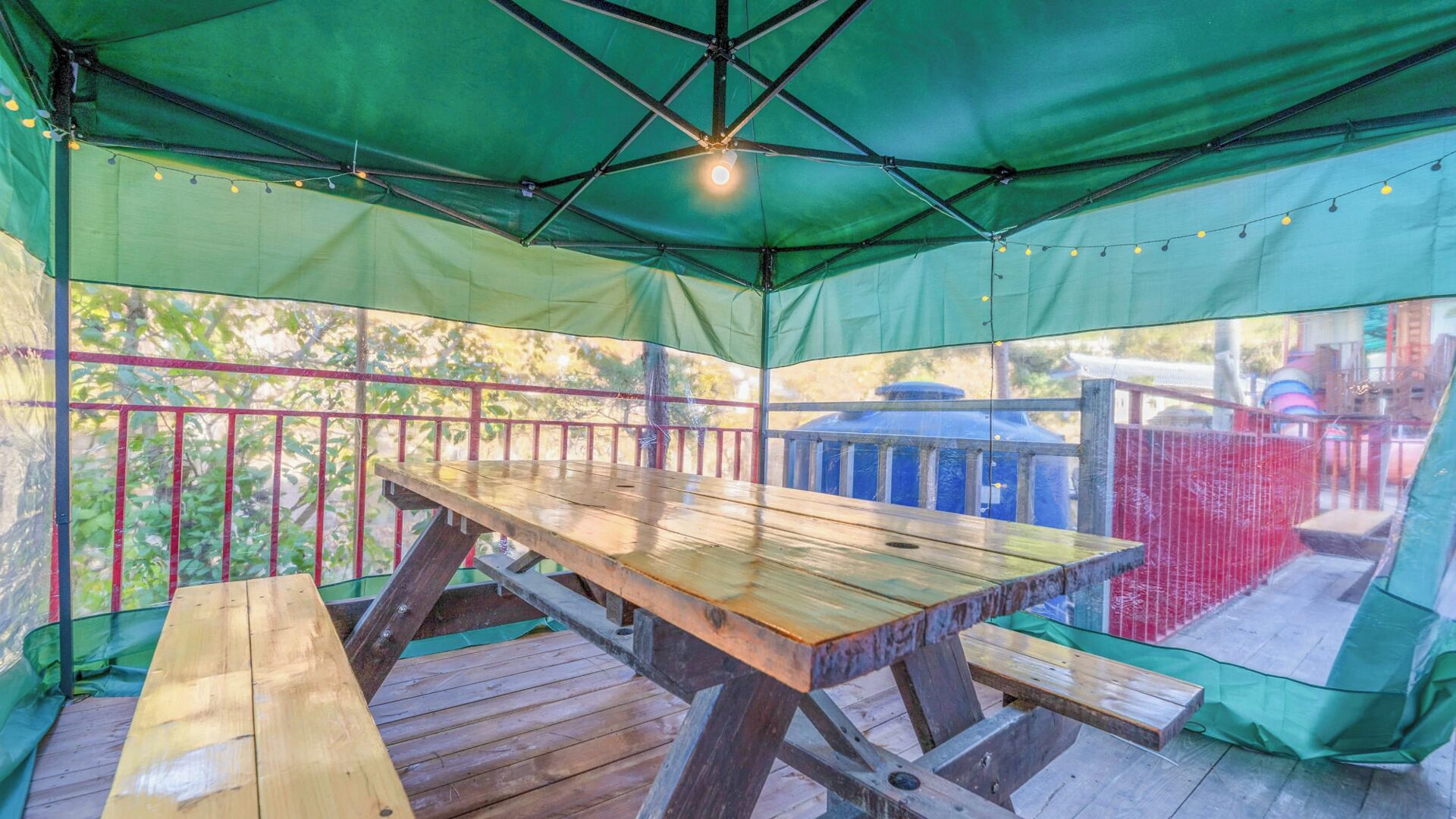 A covered outdoor seating area with a wooden table and benches, surrounded by a green canopy and string lights.