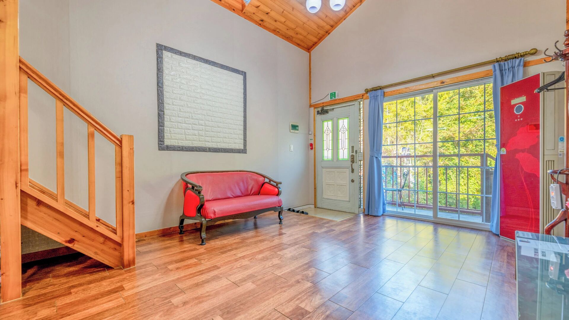A room with a wooden staircase, a red sofa, a large window with a view of greenery, and a door with a red sign.
