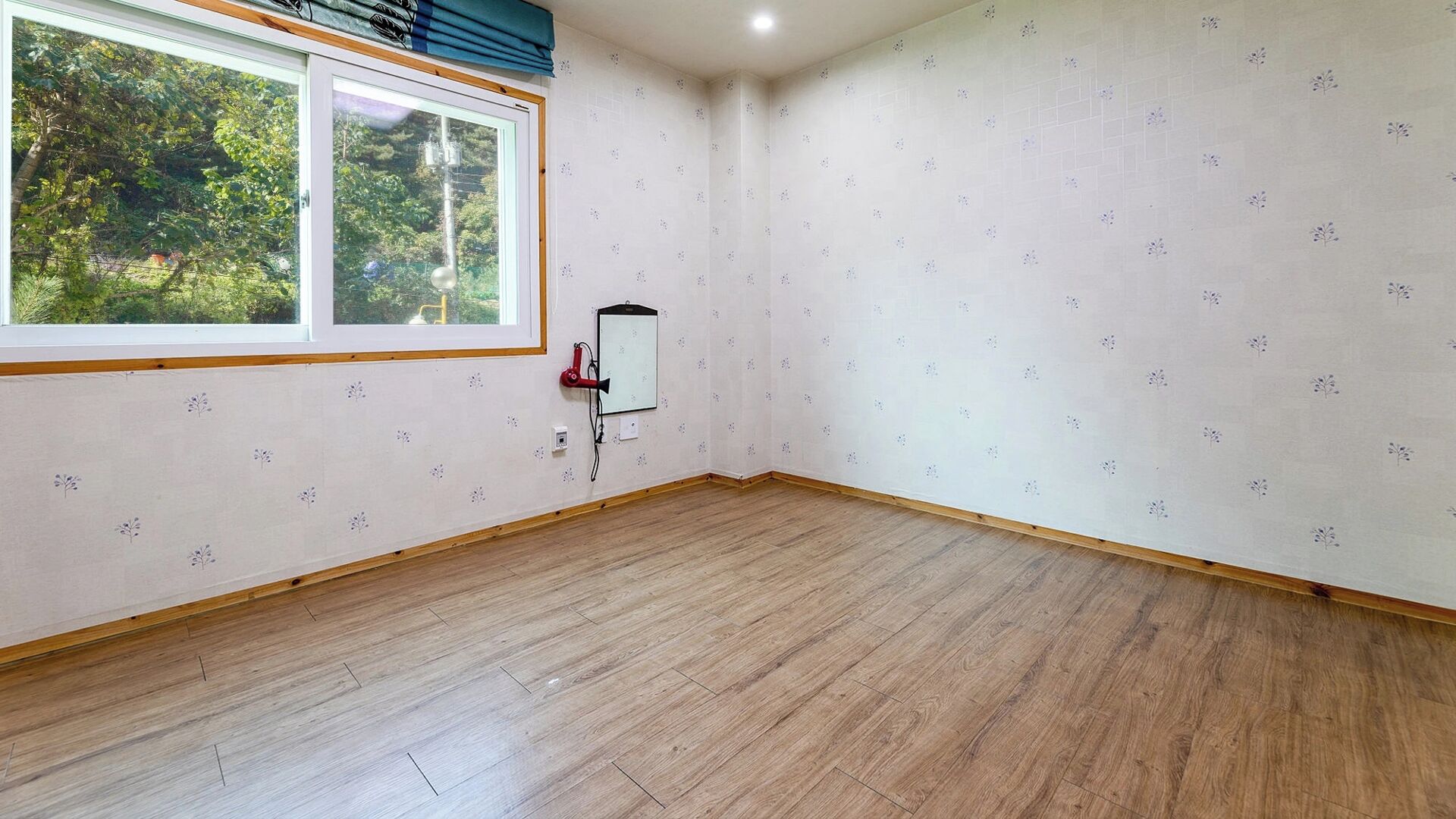 Una habitación con piso de madera, una ventana con vista a follaje y un altavoz en la pared.