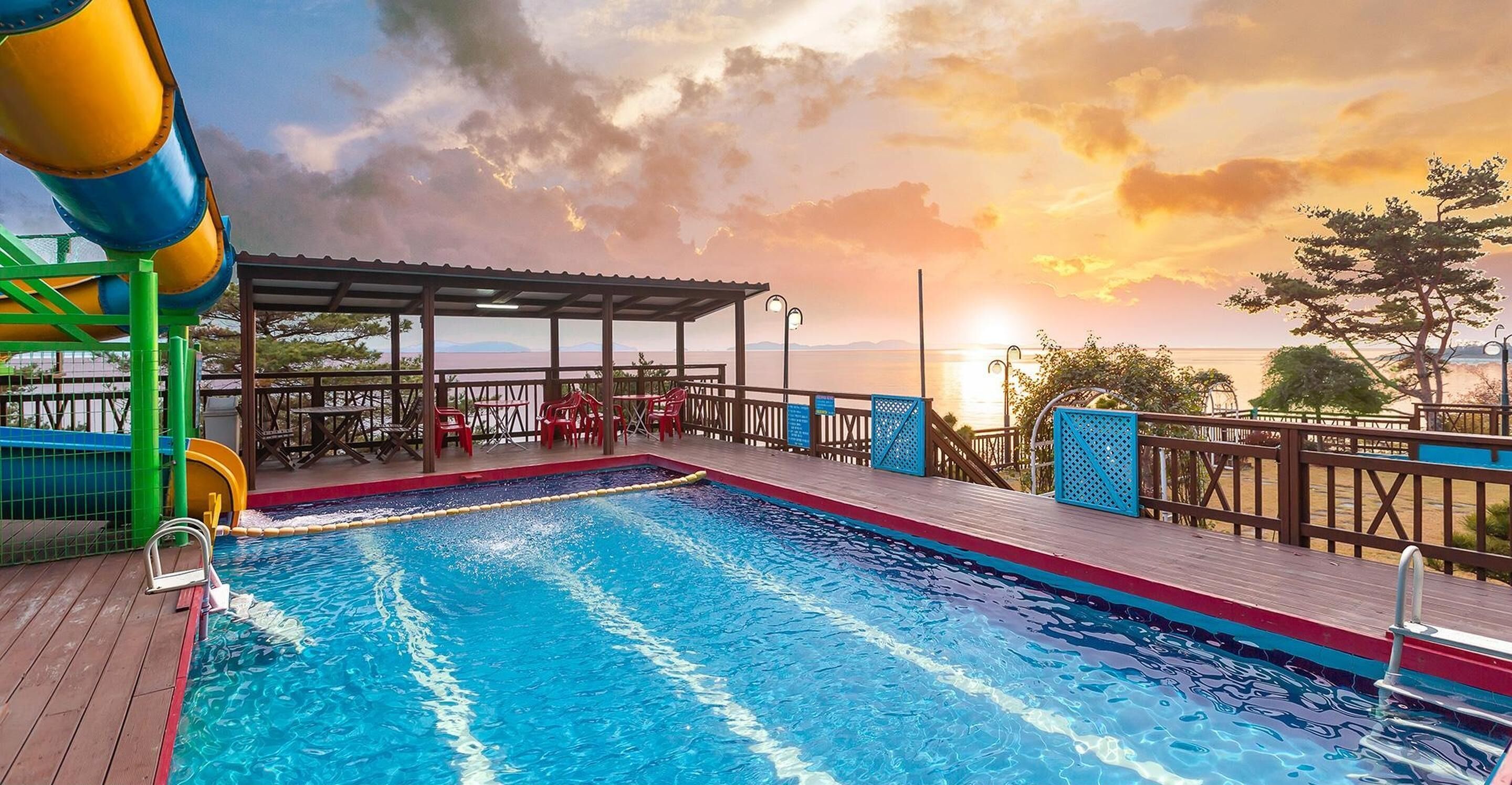 Una piscina con tobogán, un área de descanso techada y vistas al atardecer sobre el agua.
