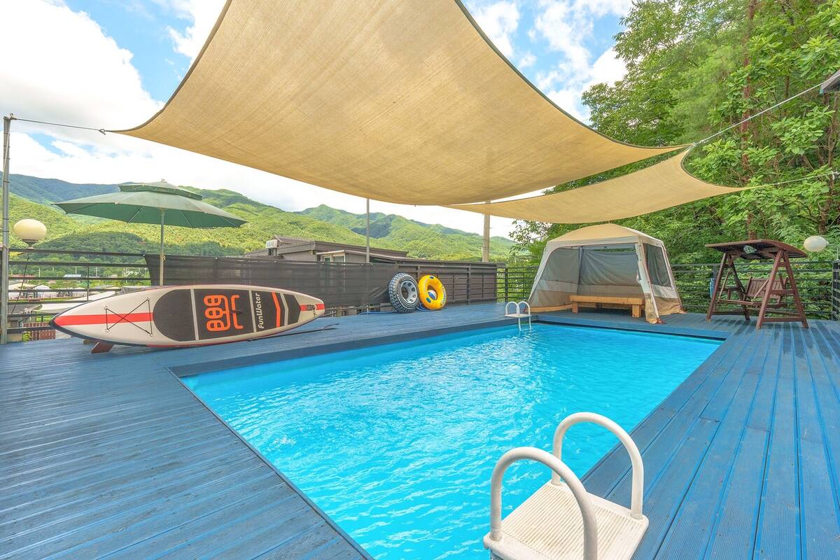 A swimming pool with a wooden deck, a tent, and a surfboard.