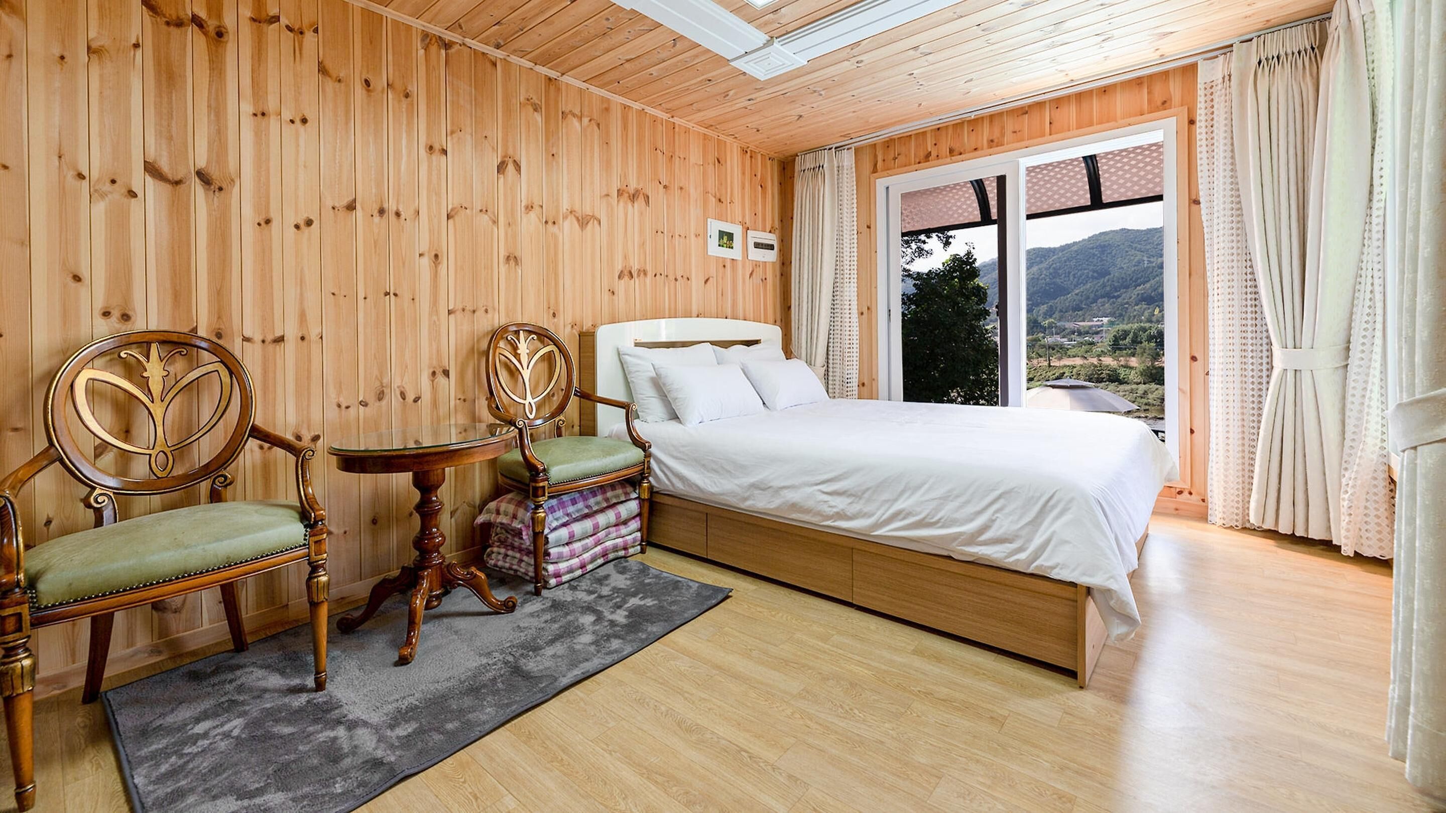 Una habitación con paneles de madera, una cama, dos sillas, una mesita y vistas a un paisaje montañoso a través de un ventanal grande.