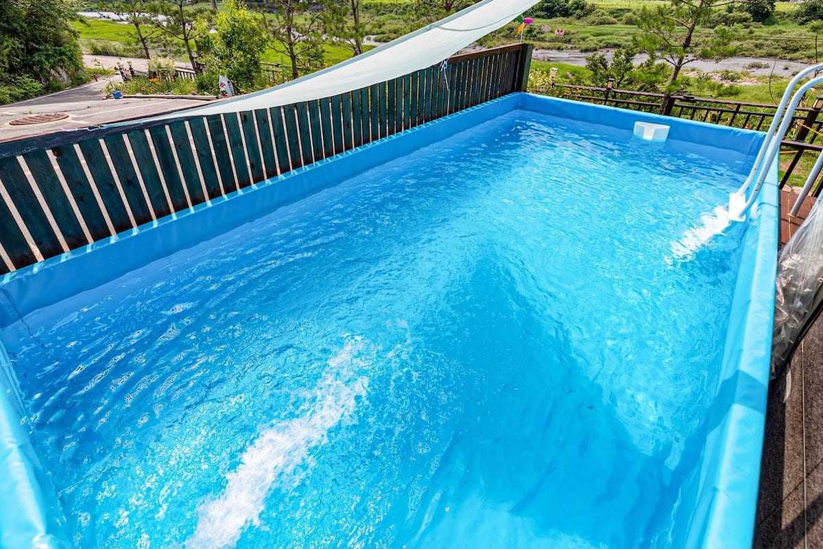 A rectangular swimming pool with a wooden deck and metal railing, surrounded by greenery.