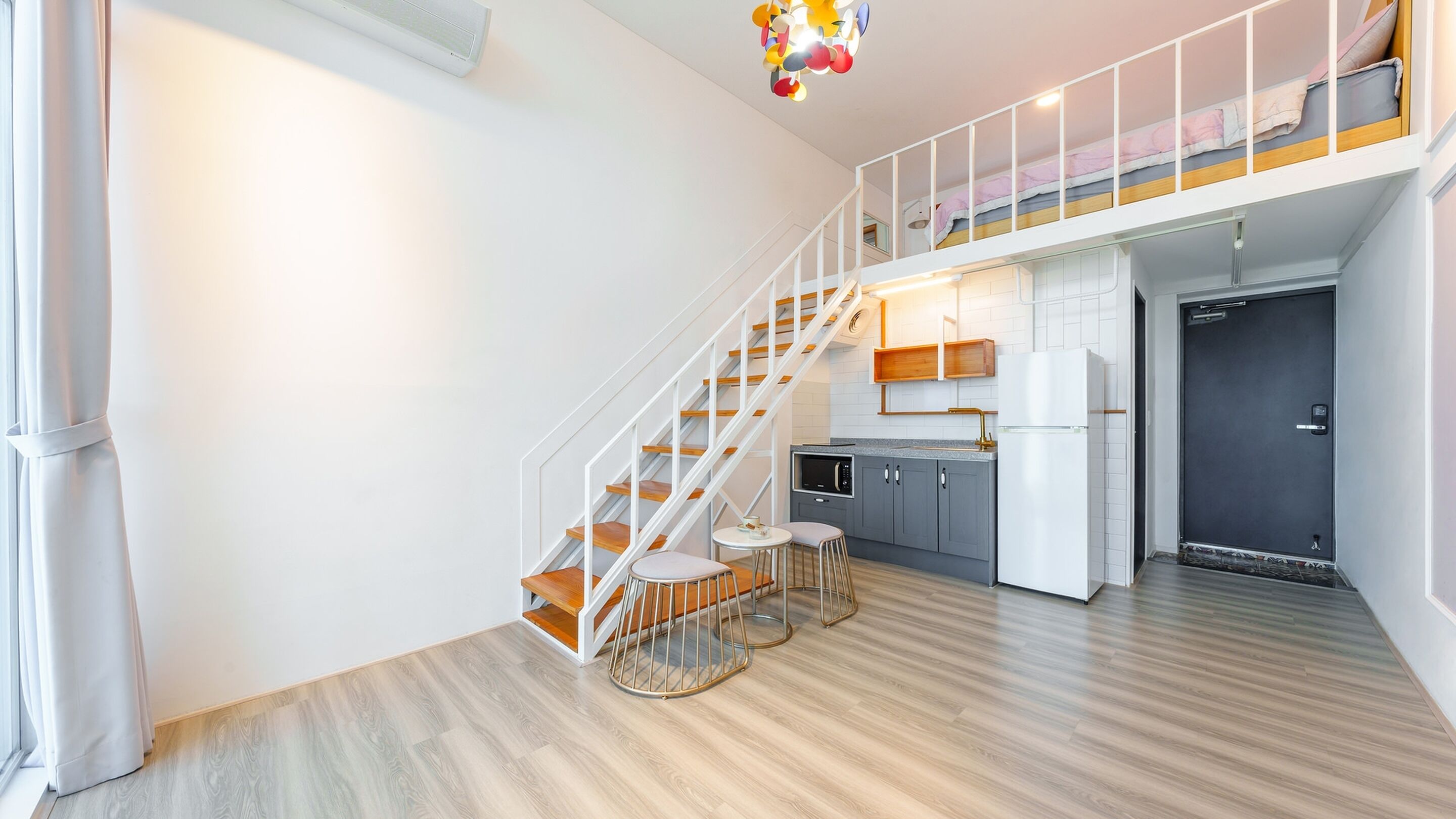 A modern apartment with a staircase leading to a loft bedroom, a kitchen with a refrigerator, and a dining area with two stools.