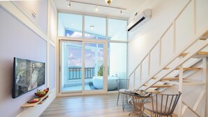 Um interior moderno com piso de madeira, uma área de refeições com mesa e cadeiras, uma TV fixada na parede e uma grande janela com vista para o exterior.