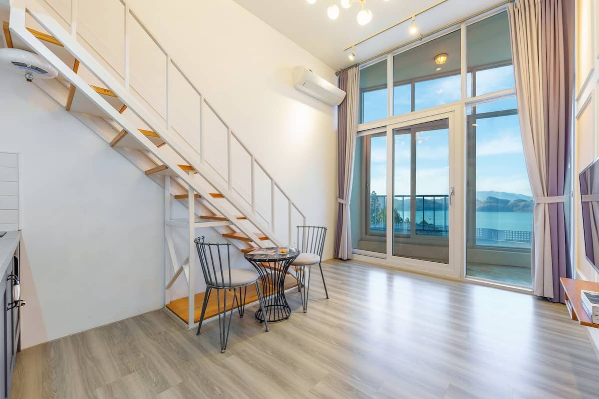 A modern apartment with a wooden staircase, a dining area with chairs, a flat-screen TV, and a large sliding glass door leading to a balcony with a view of the sea.
