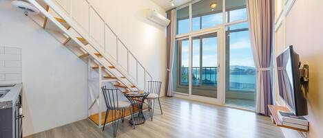 Un appartement moderne doté d’un escalier en bois, d’un coin repas avec des chaises, d’un téléviseur à écran plat et d’une grande porte-fenêtre coulissante donnant sur un balcon avec vue sur la mer.