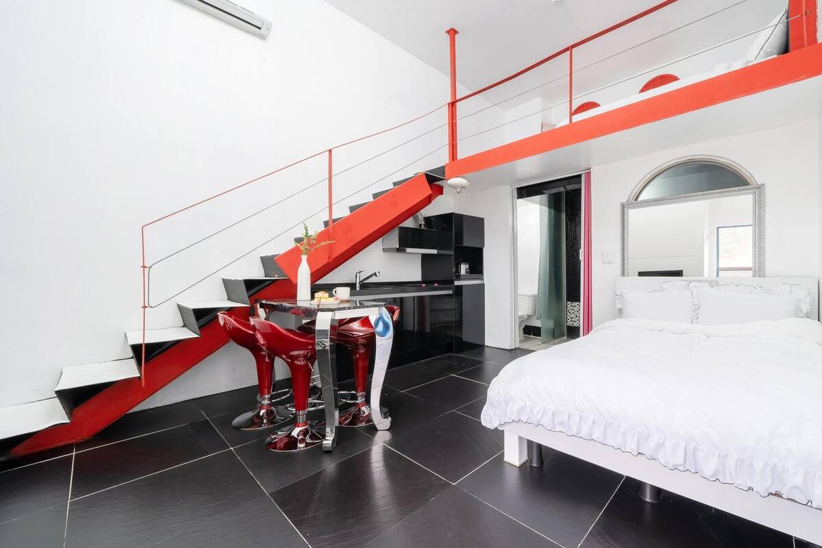 A modern bedroom with a large bed, a red staircase, and a kitchen area.