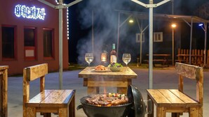 An outdoor dining setup with a grill, two wooden chairs, a table with a bottle and glasses, and a neon sign in the background. - Chuncheon Vishop Pension (Chuncheon)