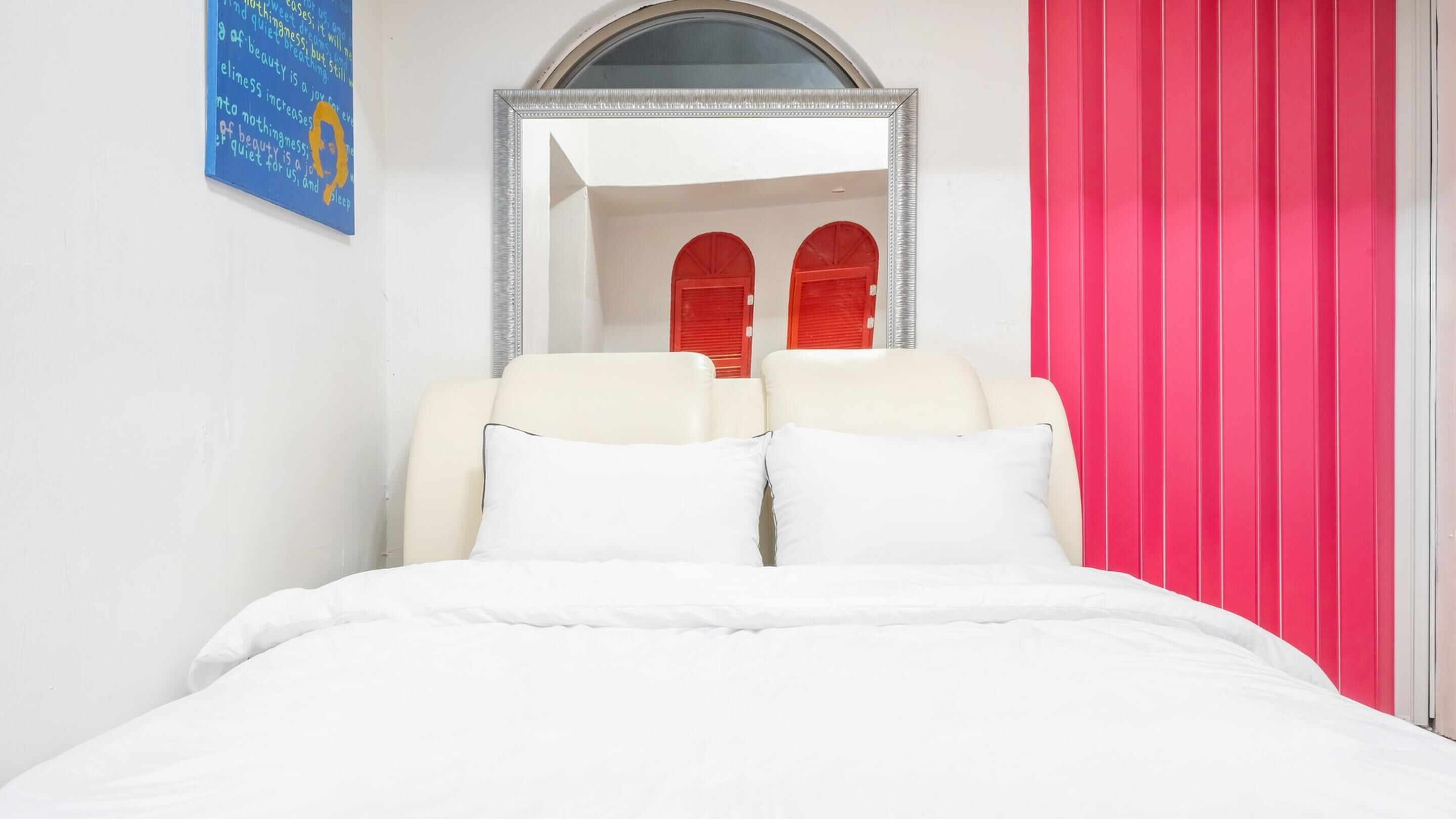 Una cama con ropa blanca, un espejo grande encima y una cortina roja a la derecha.