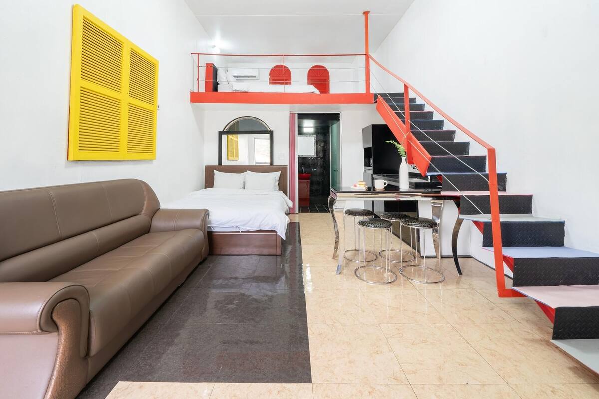 A modern hotel room with a brown leather sofa, a bed with white linens, a small kitchen area, and a red staircase.