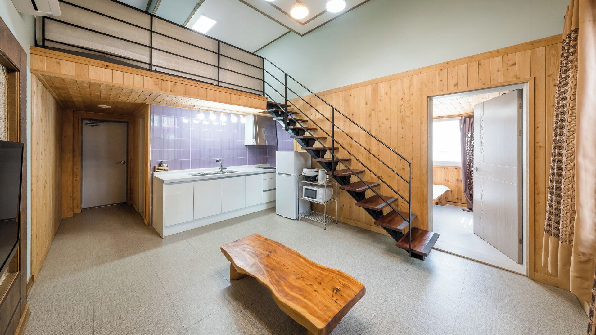 A compact kitchen with a built-in stove, a sink, and a refrigerator. There is a wooden bench and a staircase leading to an upper level.