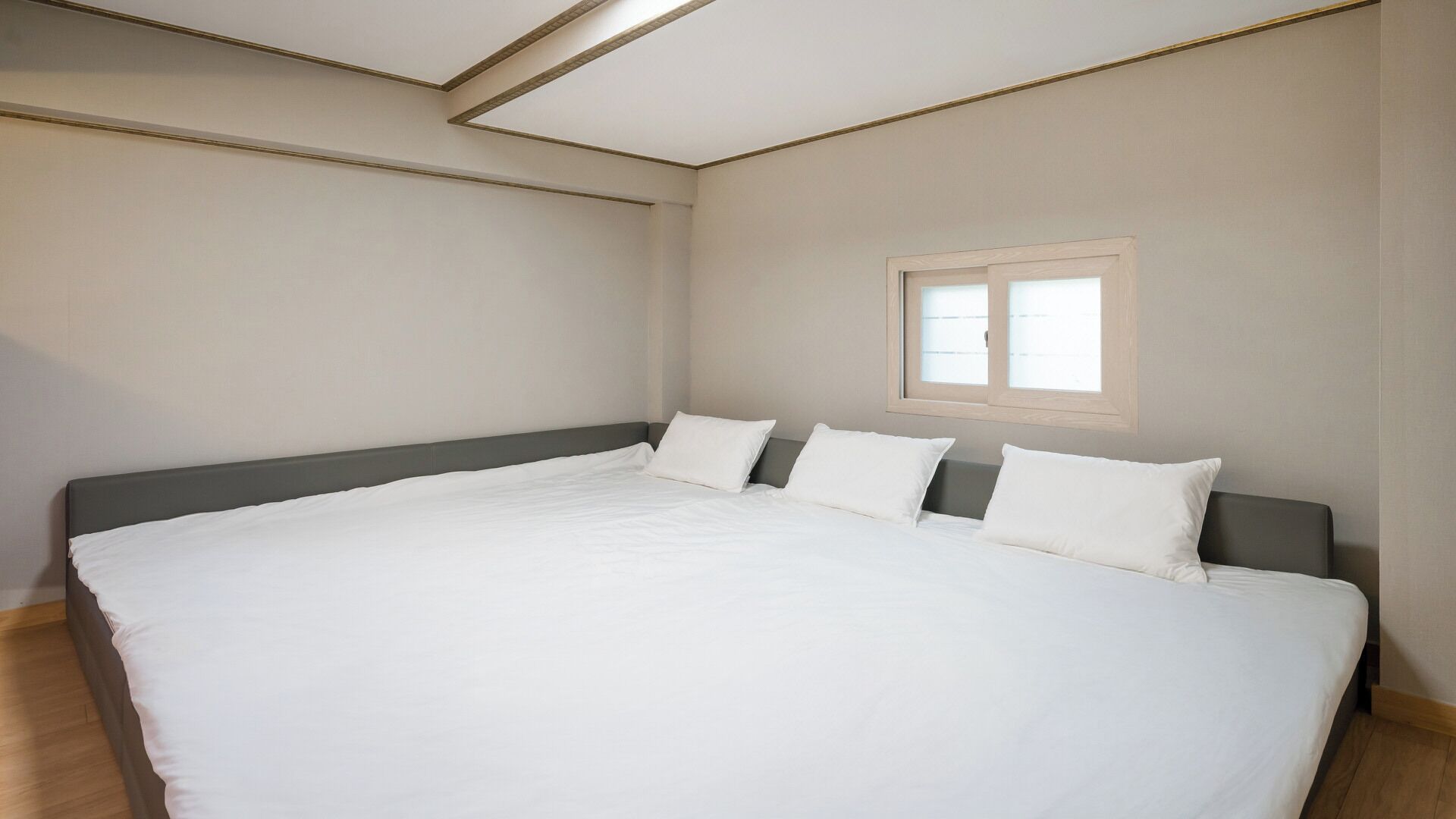 A large bed with a grey headboard, white bedding, and a small window with blinds.