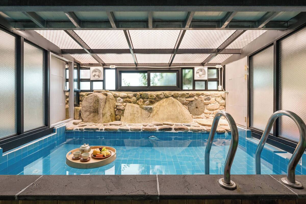 Un bain à remous avec un mur de pierres en arrière-plan, un plateau avec des fruits et du thé, et une piscine à l’eau d’un bleu limpide.