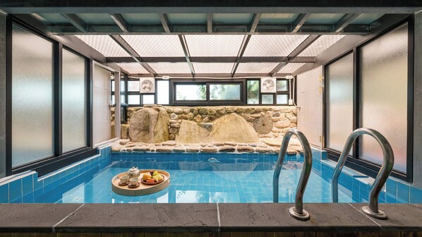 A hot tub with a stone wall backdrop, a tray with fruits and tea, and a clear blue pool.