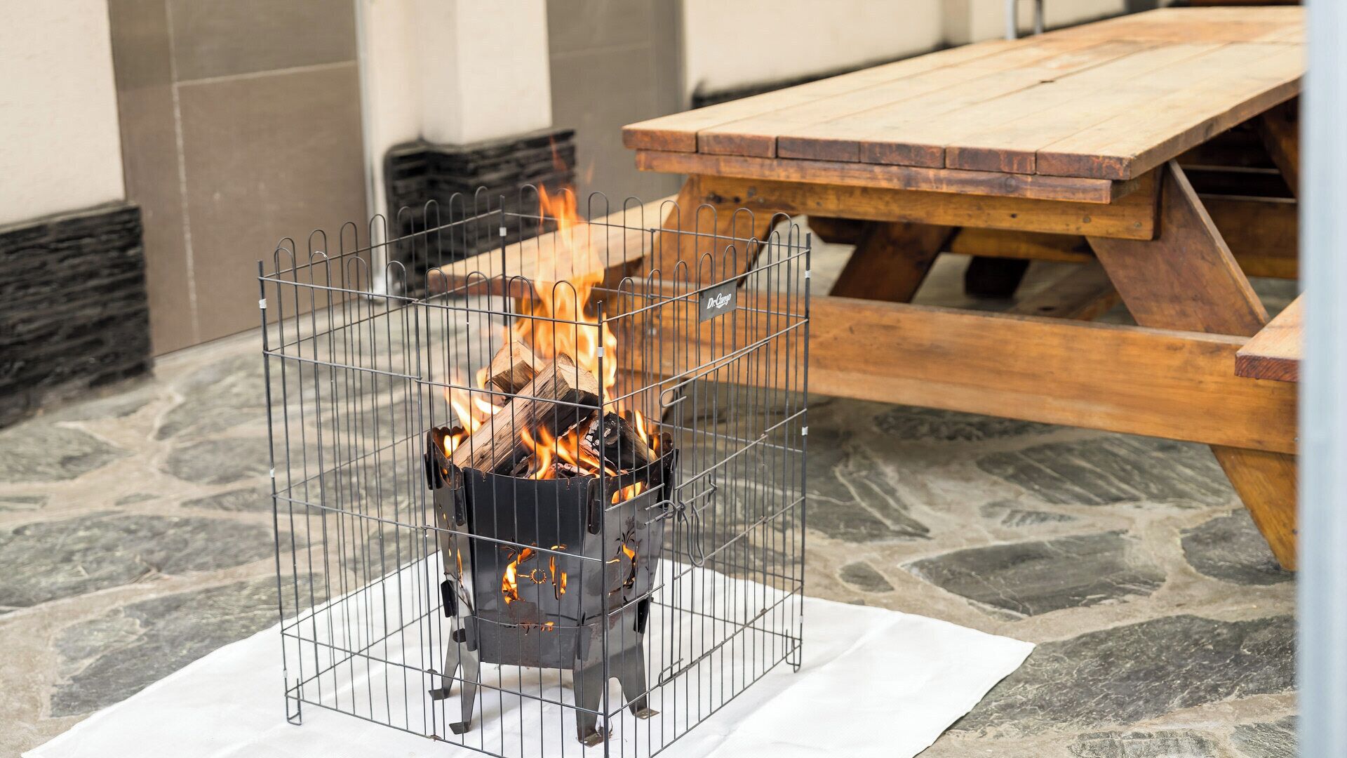 A metal fire pit with burning logs, a wooden picnic table, and stone flooring.