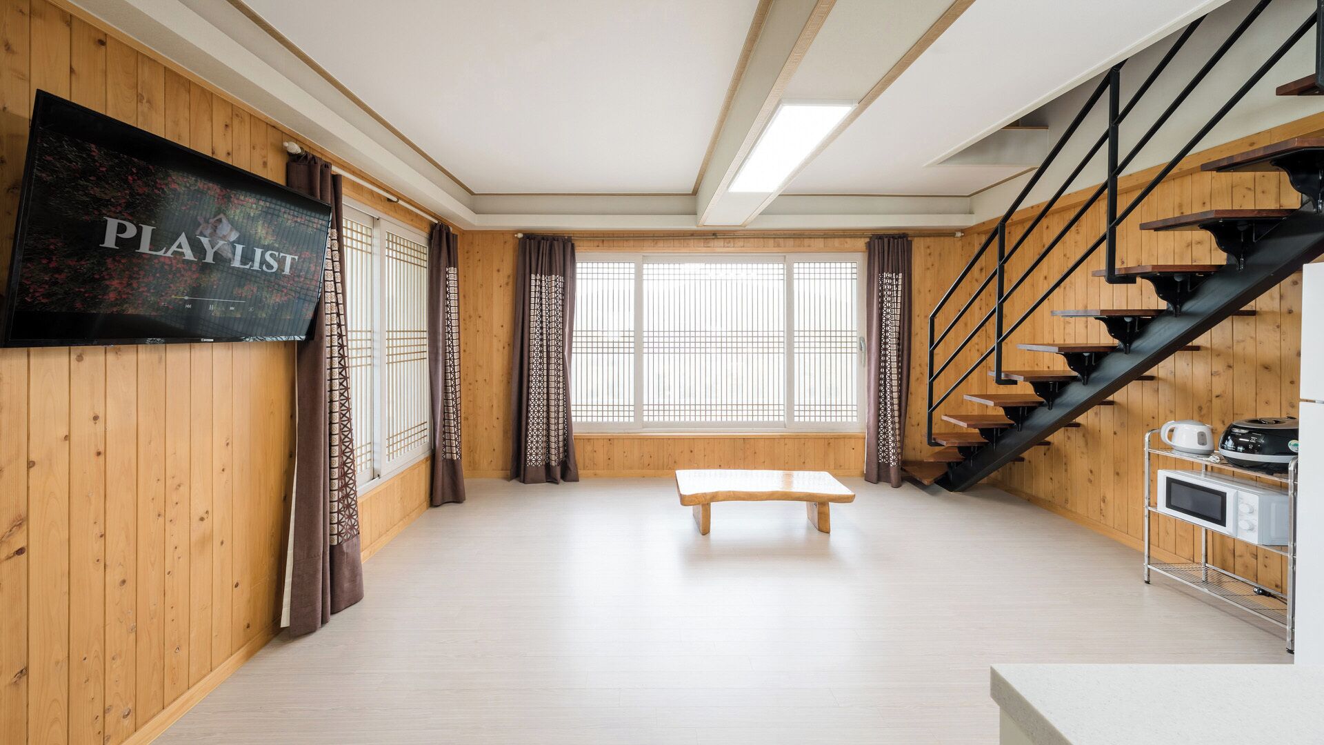 A room with a wooden floor, a bench, a staircase, a TV, and a window with curtains.