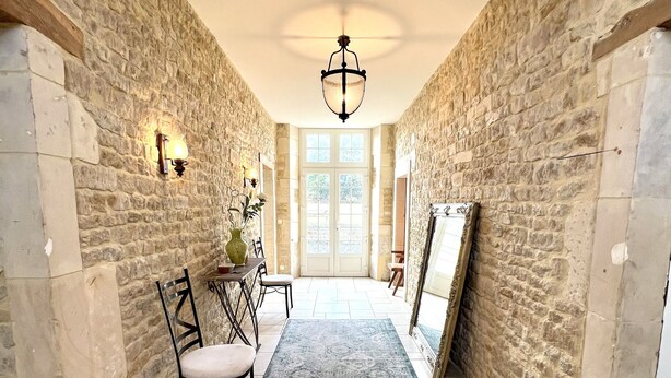 A stone-walled hallway with a dining area, a chandelier, and a large window at the end.