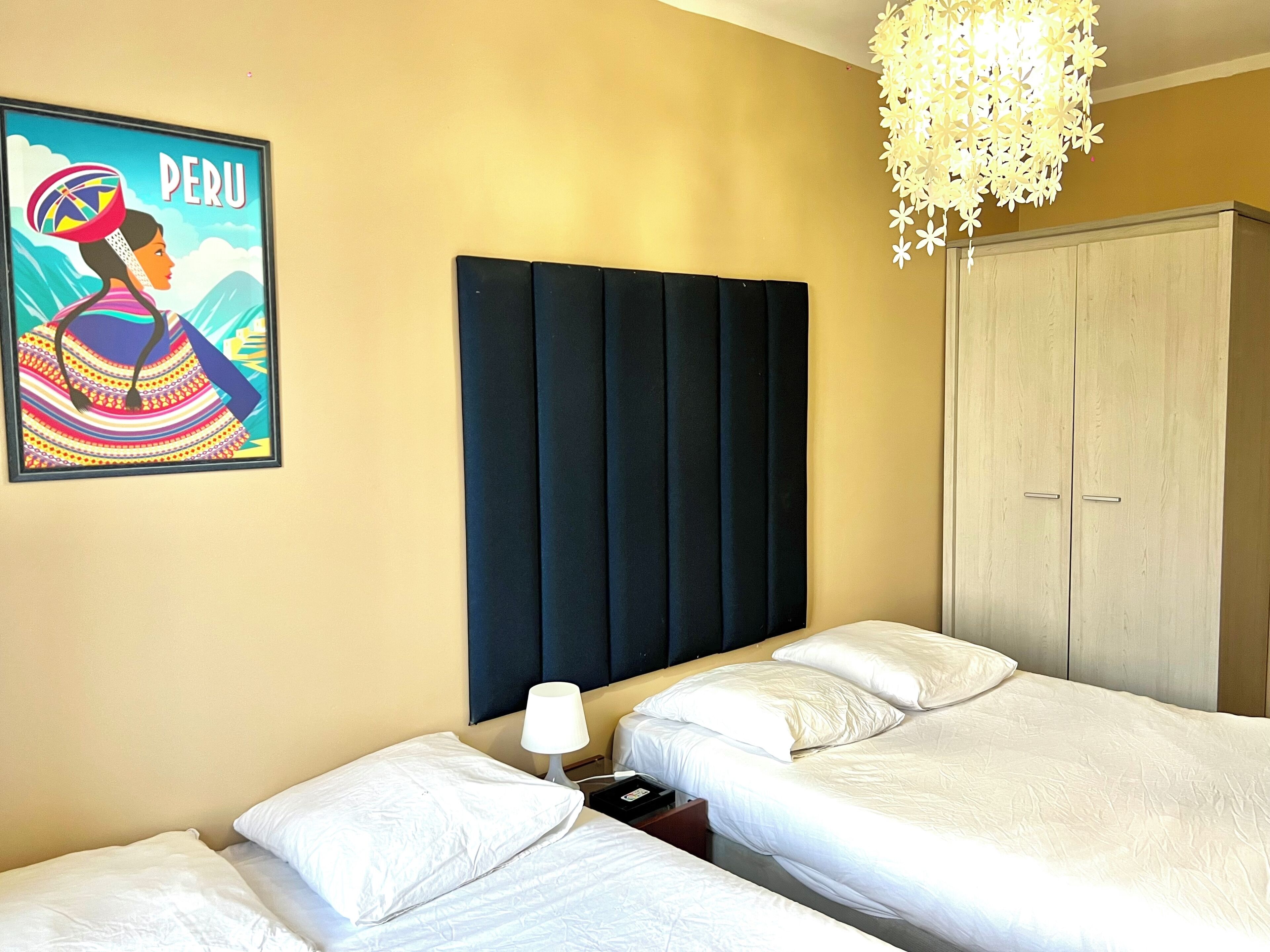 Een hotelkamer met twee bedden, een grote kledingkast, een vastgeschroefd hoofdbord en een poster van een Peruaanse vrouw.