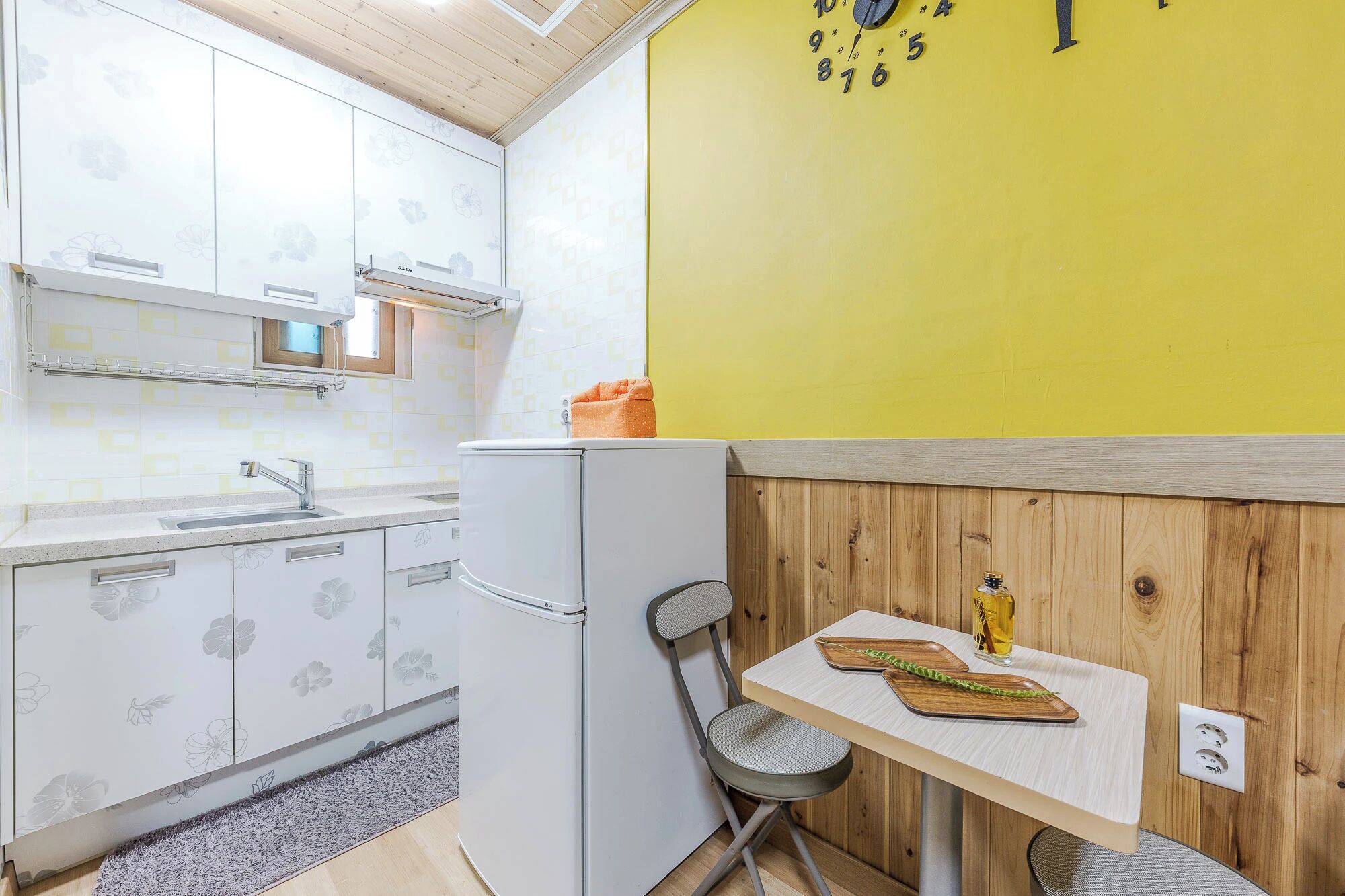 Een compacte keuken met een witte koelkast, een gootsteen en een houten wand met een gele klok.