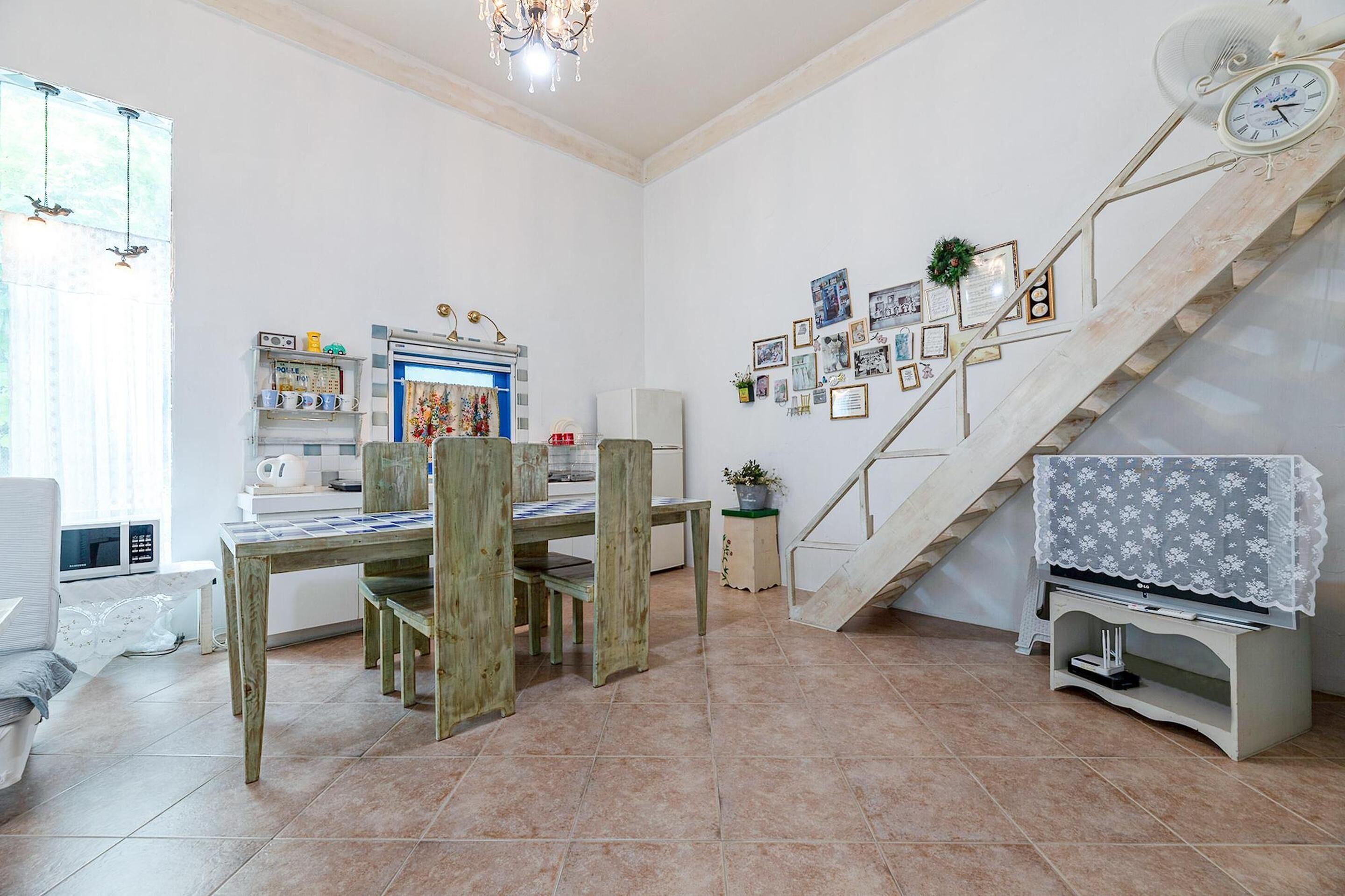 Un comedor con mesa y sillas de madera, una zona de cocina con electrodomésticos y una escalera con un reloj y fotografías enmarcadas.