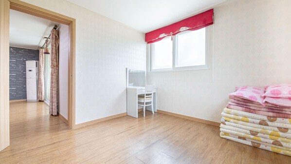 Una habitación con piso de madera, un escritorio blanco con una silla, un ventanal con cortina roja y una pila de toallas dobladas.
