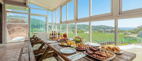 A long wooden table set for a meal with various dishes, bottles, and a scenic view through large windows.