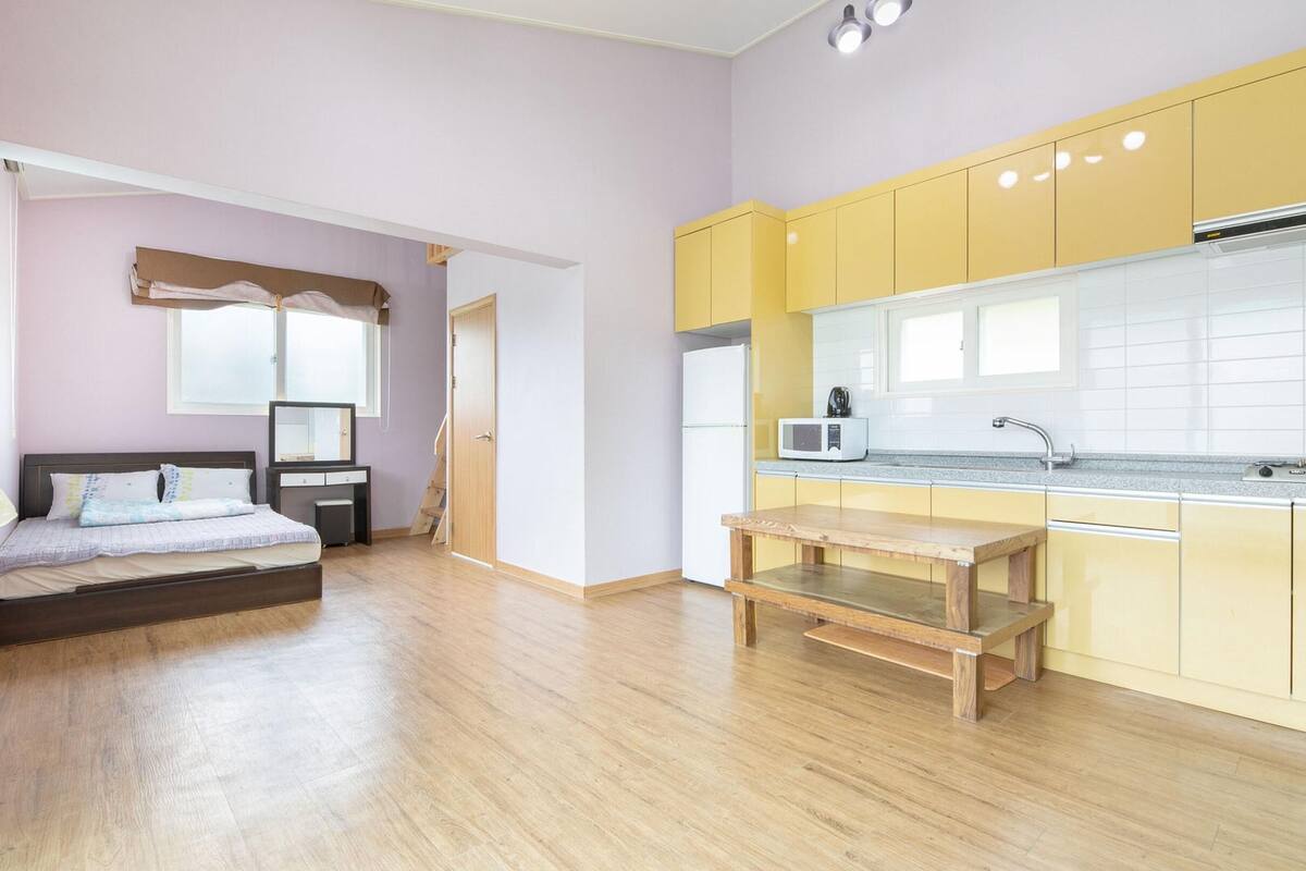 A compact room with a bed, a small desk, a bench, a kitchenette, and a window.
