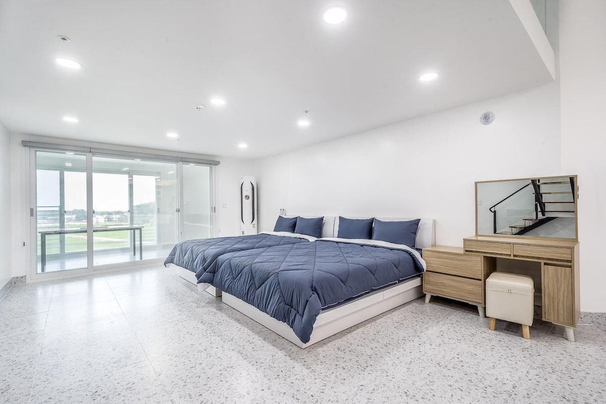 A modern bedroom with a large bed, a wooden dresser, a mirror, and a balcony with a view.