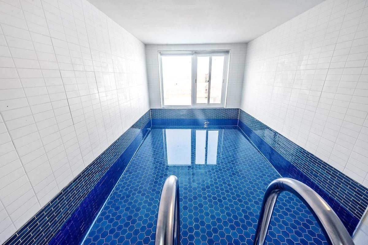 A swimming pool with blue hexagonal tiles and metal handrails, surrounded by white tiled walls and a window at the far end.