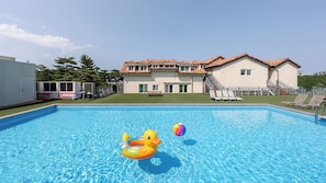 A swimming pool with inflatable toys, a patio with lounge chairs, and a building with a tiled roof in the background.