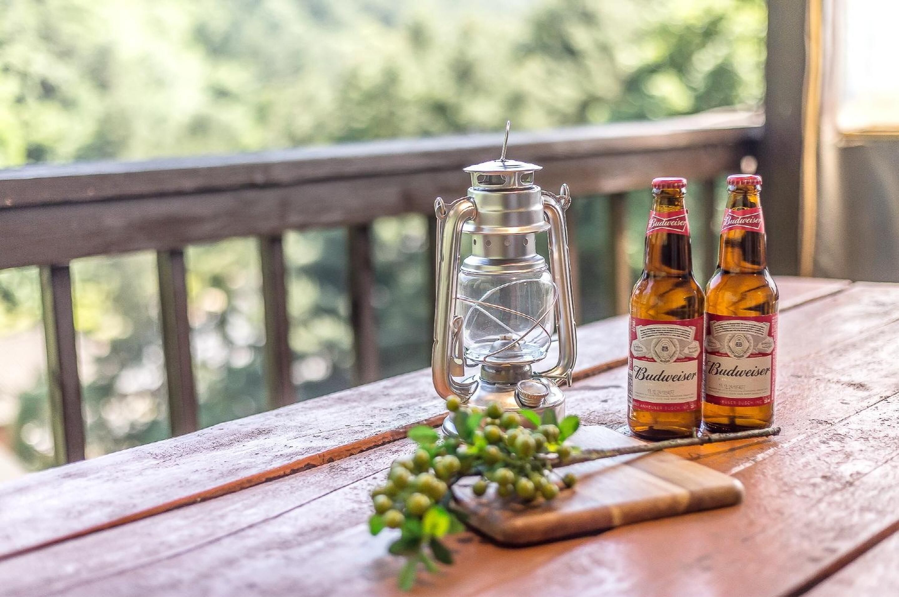 A wooden table with two Budweiser beer bottles and a clear glass lantern on a balcony with a view of trees.