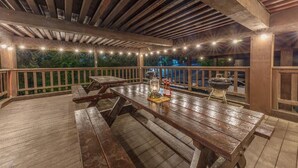 Eine Terrasse im Freien mit Holzbänken und -tischen, Lichterketten und einem Grill.