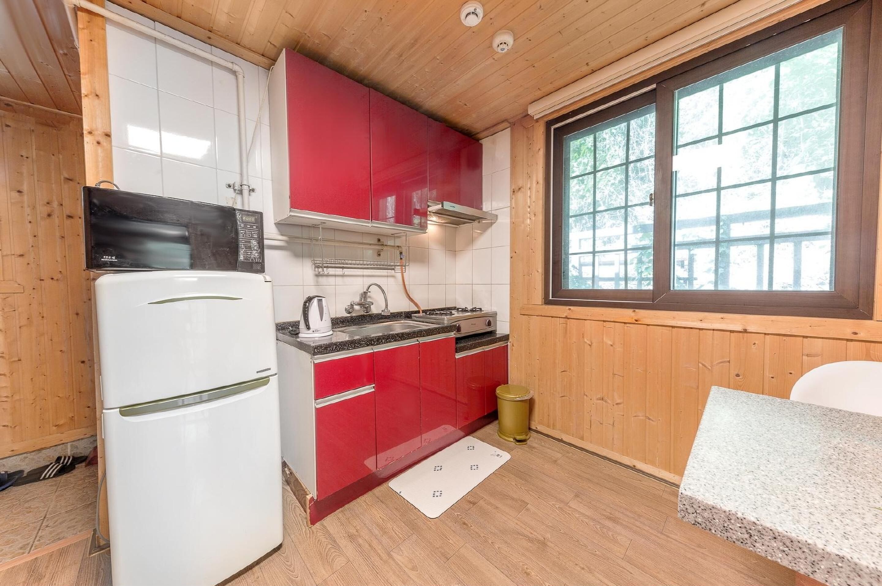 A compact kitchen with a white refrigerator, red cabinets, a stove, and a window with a view of trees.