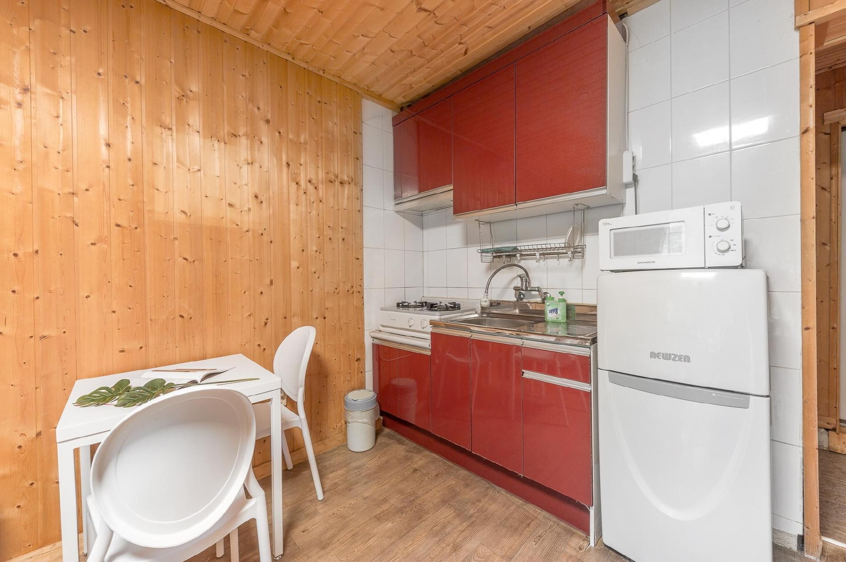 Een kleine keuken met houten wanden, een witte tafel met stoelen, een magnetron en een rode koelkast.