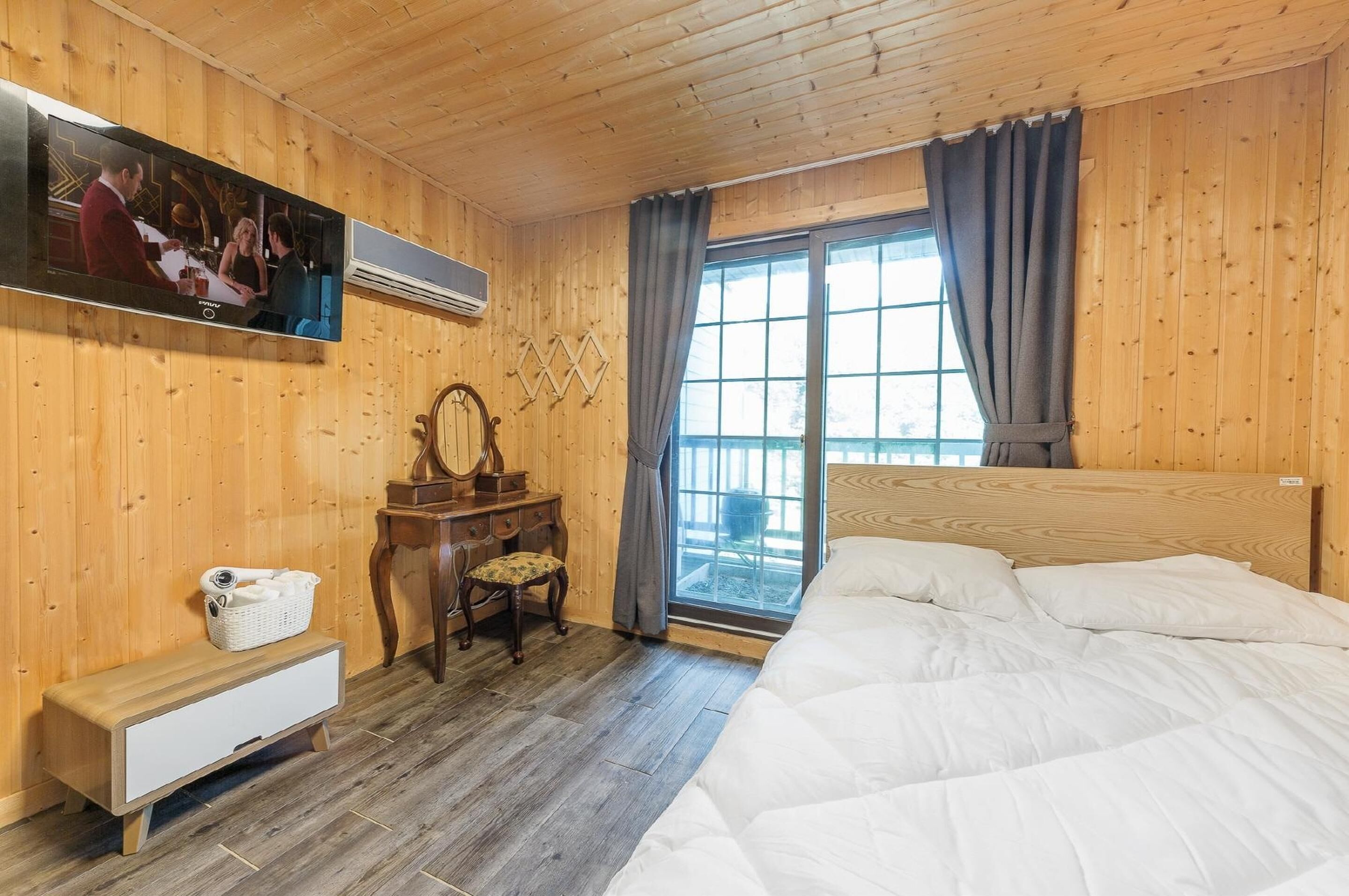 Ein Zimmer mit Holzwandverkleidung, das ein Bett, einen Schreibtisch, einen Stuhl, einen Fernseher und ein Fenster mit Vorhängen beinhaltet.
