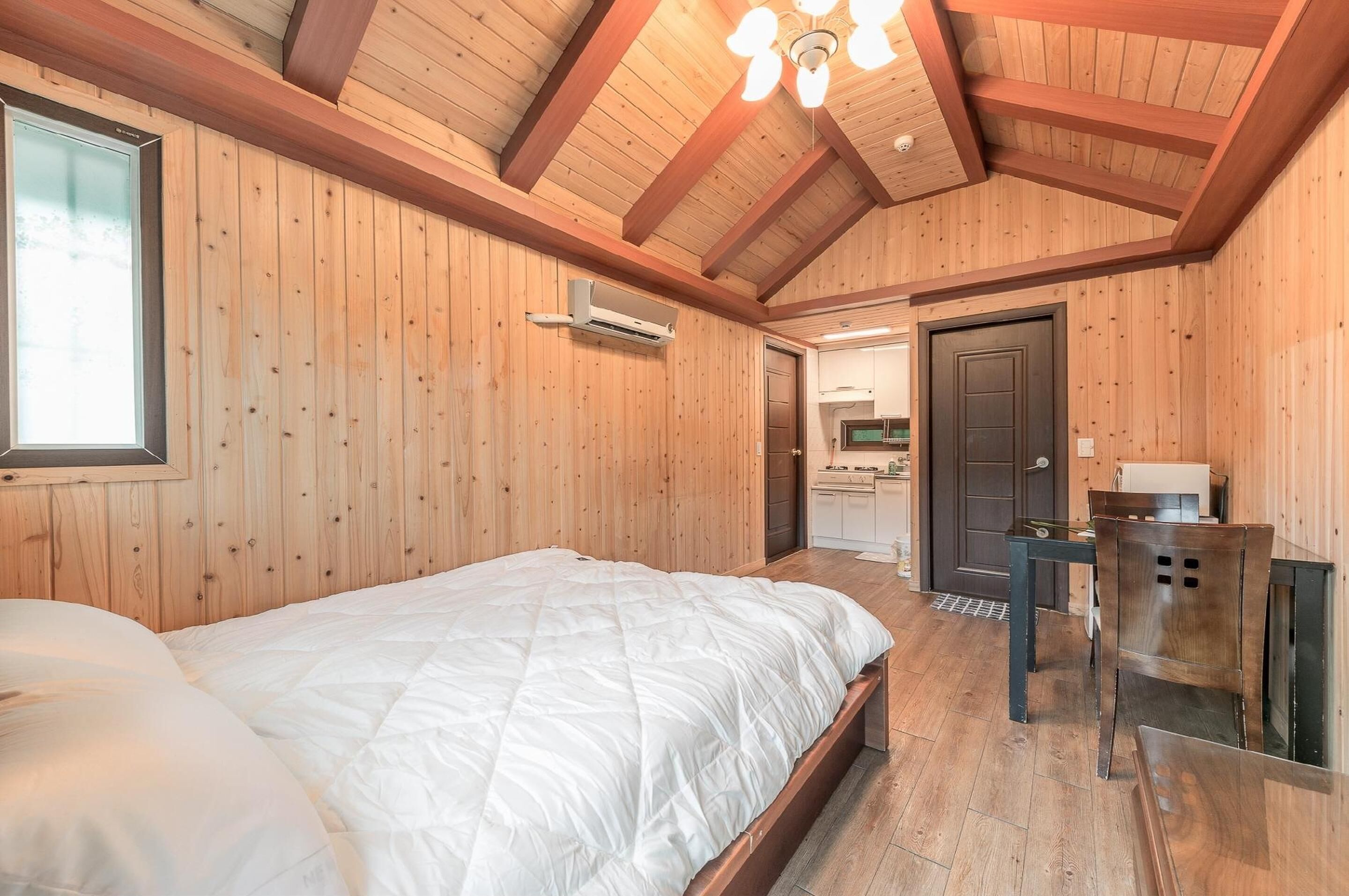 A wooden-paneled room with a bed, a table, and a door leading to a kitchen.