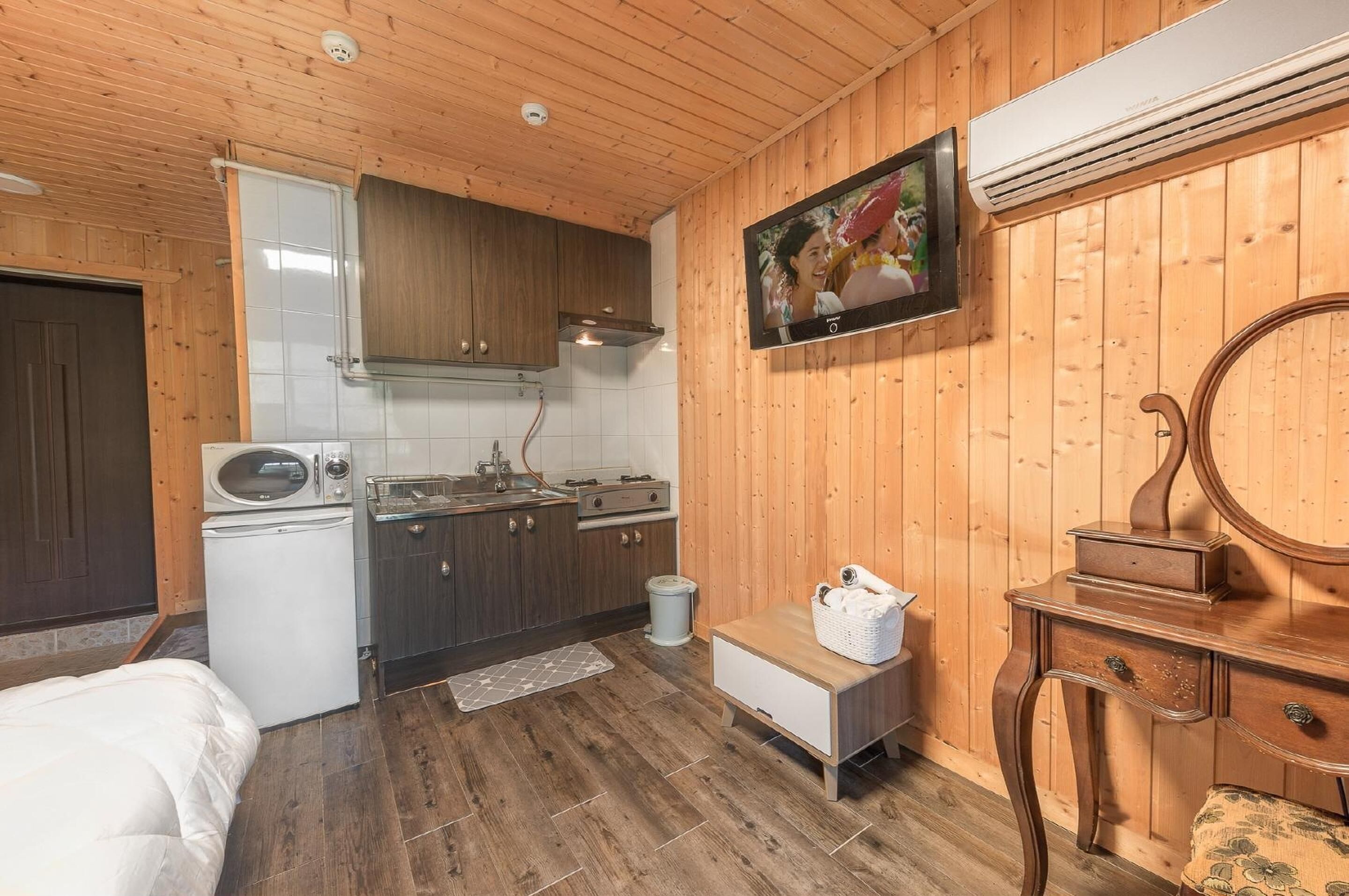 A compact room with wooden walls, a kitchenette, a bed, a small table with a basket, and a mirror.