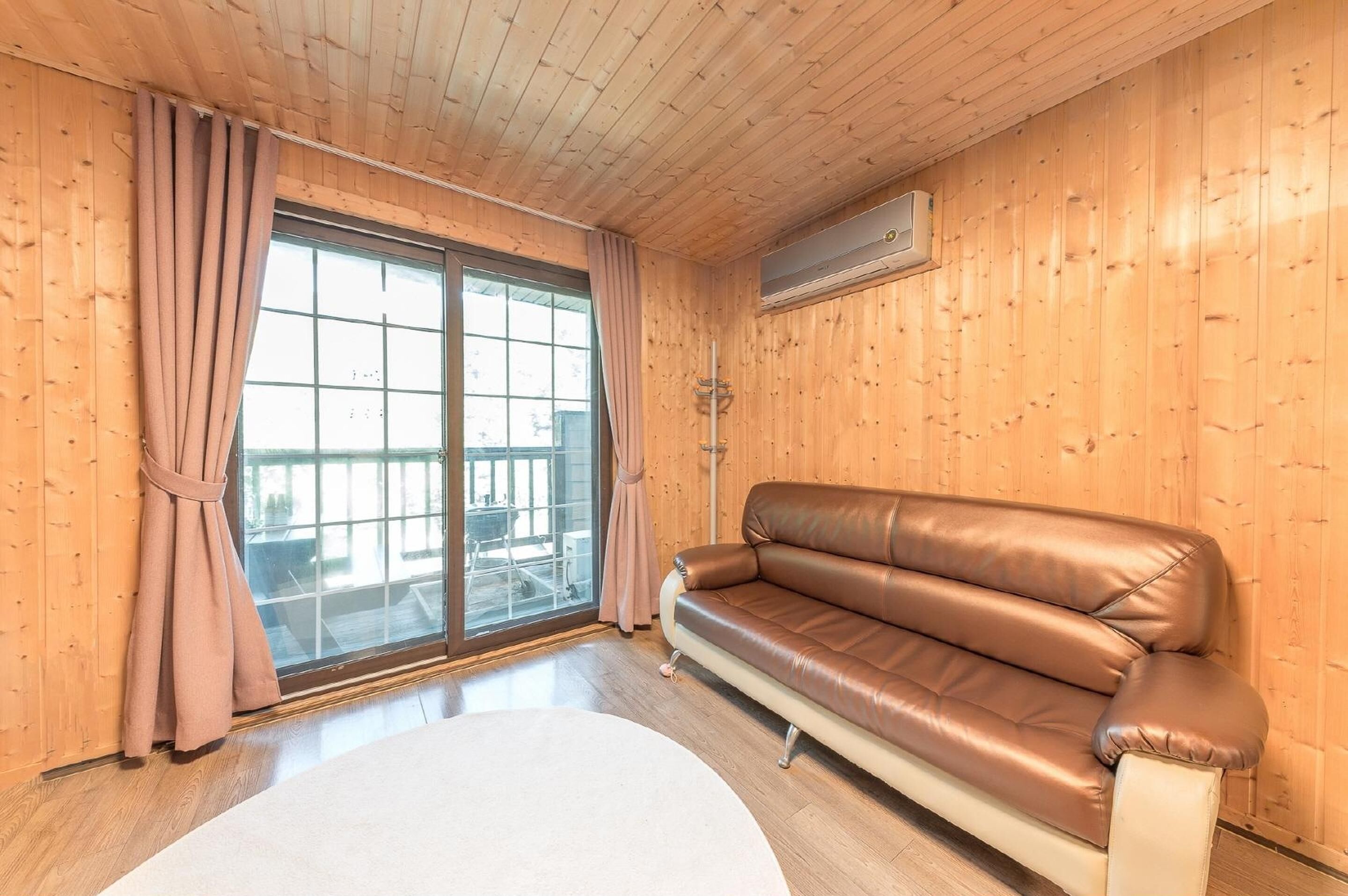 A wooden-paneled room with a brown leather sofa, a large window with curtains, and a white round table.
