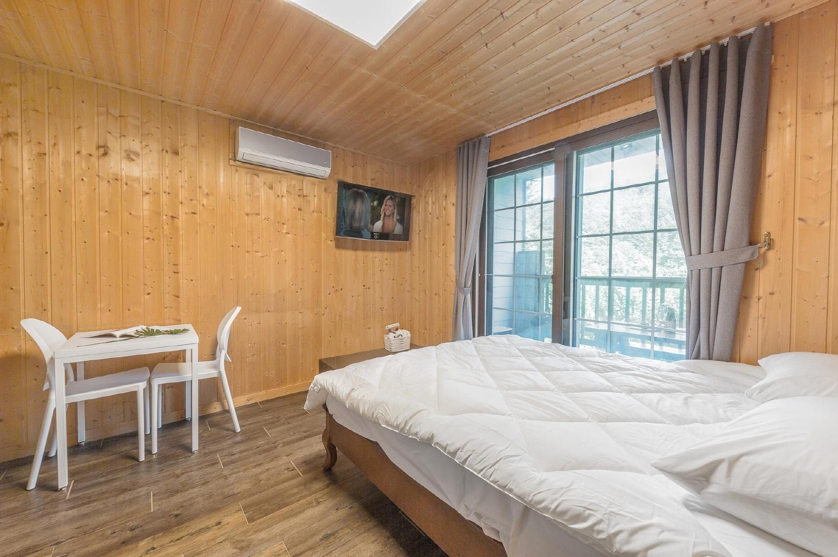 Habitación con paneles de madera, una cama, una mesa con sillas y un ventanal con cortinas.
