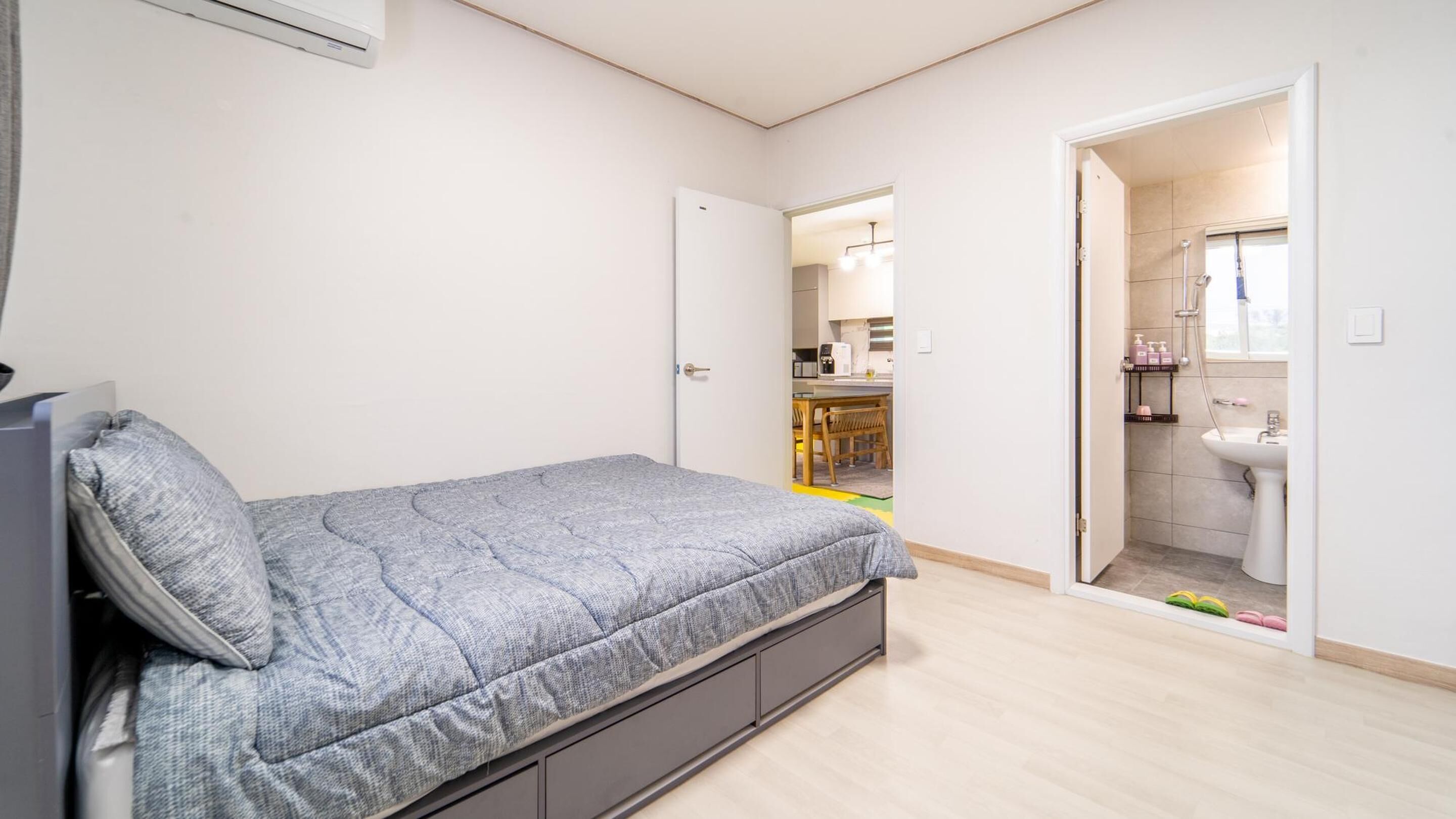 A bedroom with a bed, a door leading to a bathroom, and a kitchenette in the background.